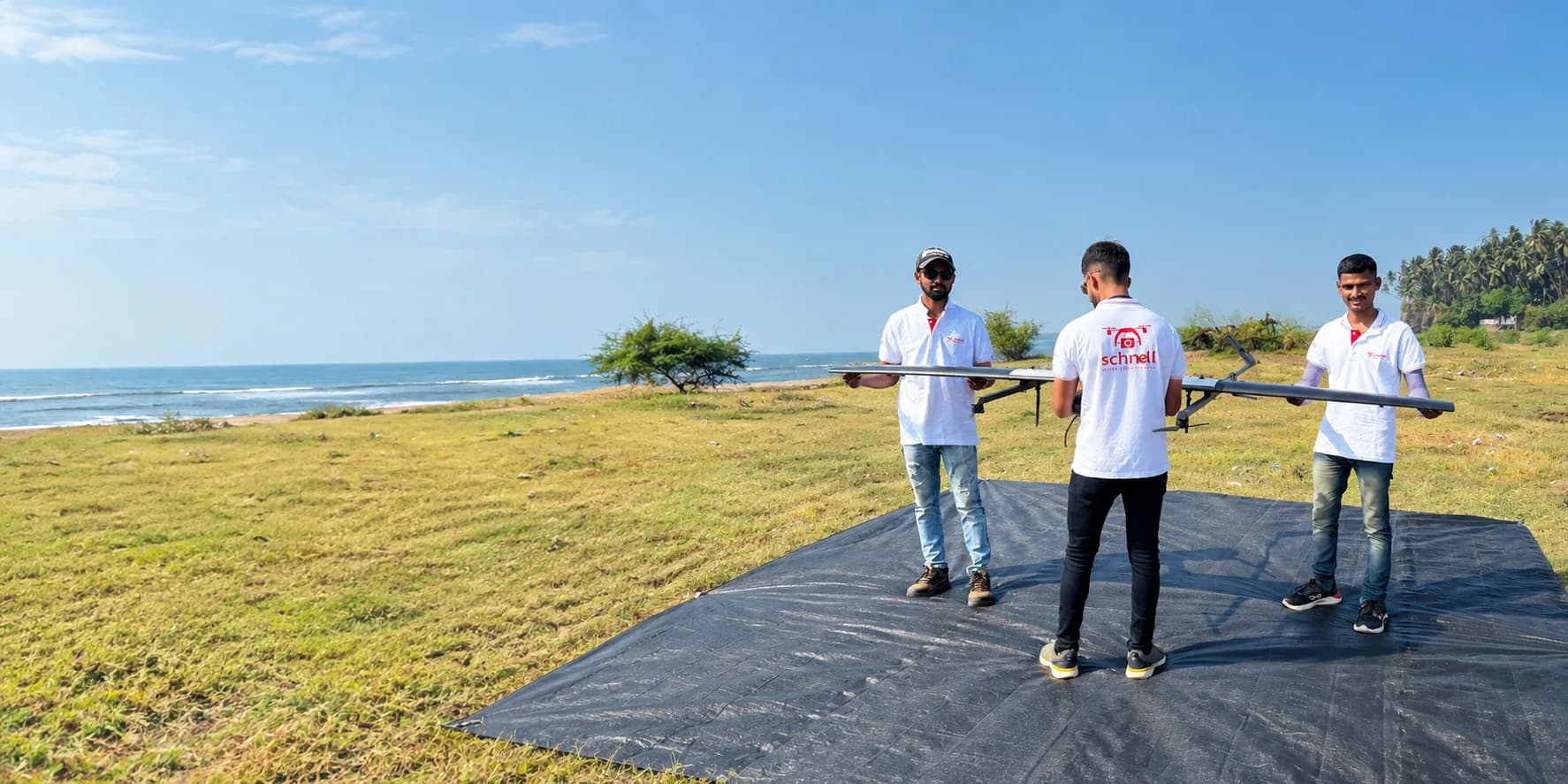 Three Schnell drone pilots carrying a fixed-wing survey drone onto a launch mat at a coastal site