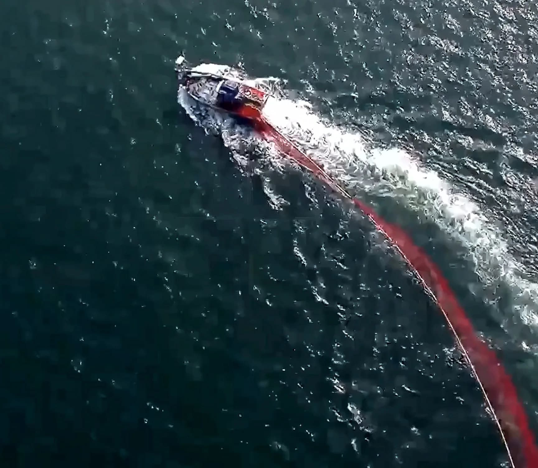 A fishing vessel under way in dark open water, a long red net trailing behind it